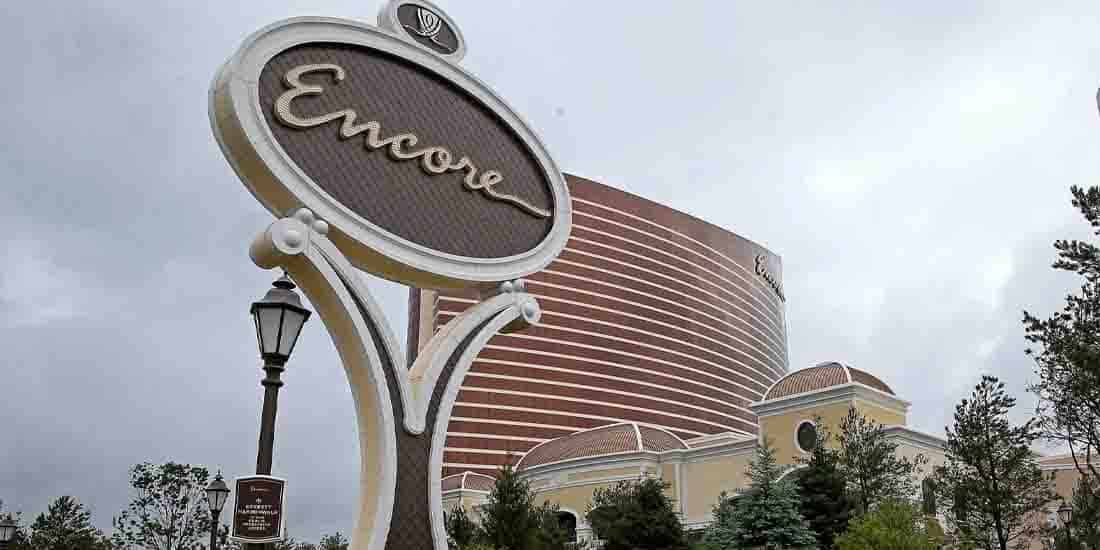 Large retro-style 'Encore' sign on a Y-shaped pedestal in front of a curved beige hotel building with trees and a lamppost nearby, daytime cloudy sky.