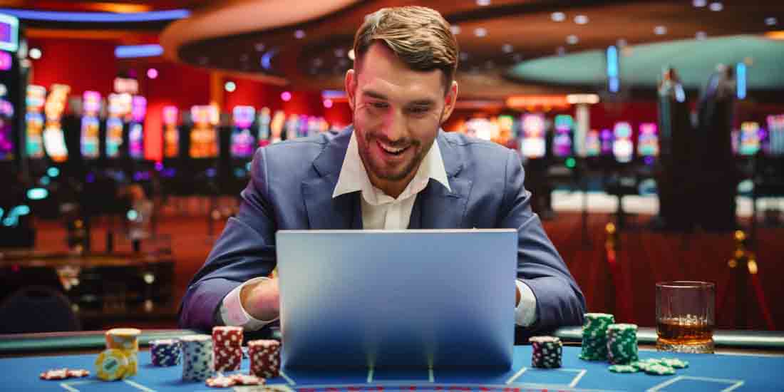 Man in a blue suit using a laptop at a casino table, surrounded by poker chips and a glass of whiskey, with colorful slot machines in the background.